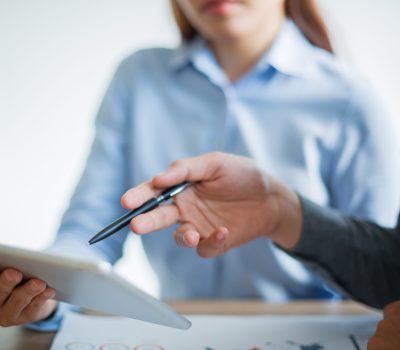 Unrecognizable Asian business team having meeting in office. Businesswoman holding touchpad, businessman pointing at touchscreen with pen and discussing information
